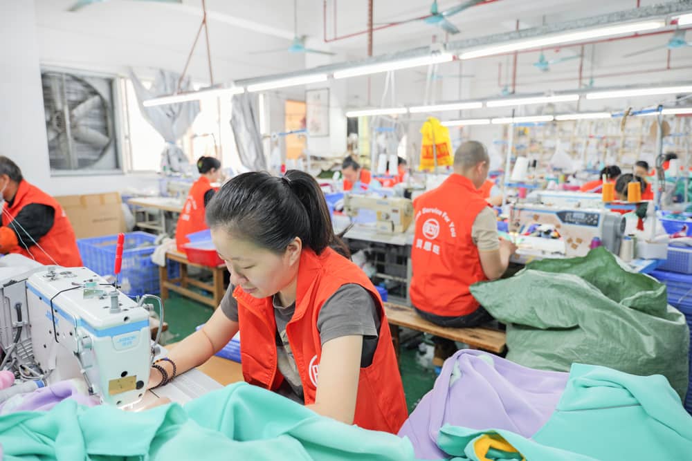 Workers in red vests using sewing machines in a busy manufacturing workshop.