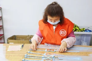 Рабочий в оранжевом жилете с надписью 'BEST' собирает продукцию на столе на фабрике.