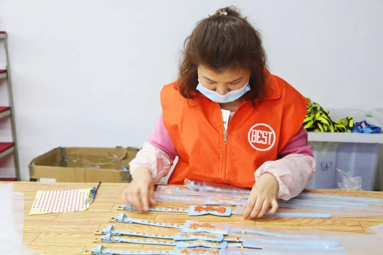 Worker in an orange vest labeled 'BEST' assembling products on a table in a factory setting.