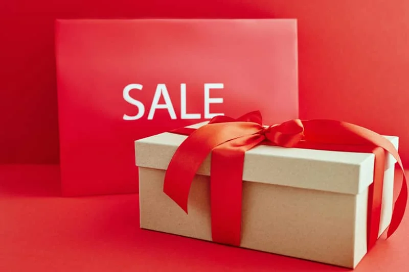 Gift box with red ribbon in front of a red sign with the word 'SALE' on a red background.