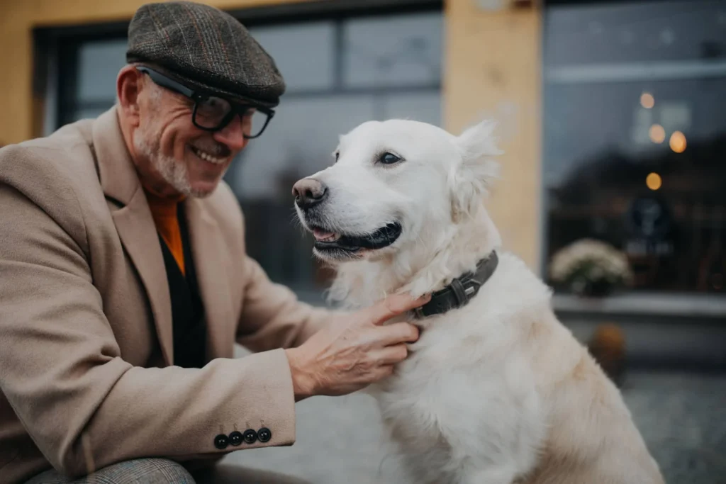 A man in a brown coat and flat cap gently petting a happy white dog outside a building.