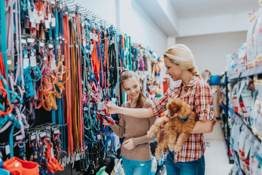 Two women selecting dog leashes in a pet store aisle, one holding a small brown dog.